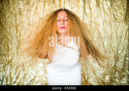 Une jeune femme aux longs cheveux blonds blonde vêtue d'une robe blanche en face d'une toile d'or Banque D'Images