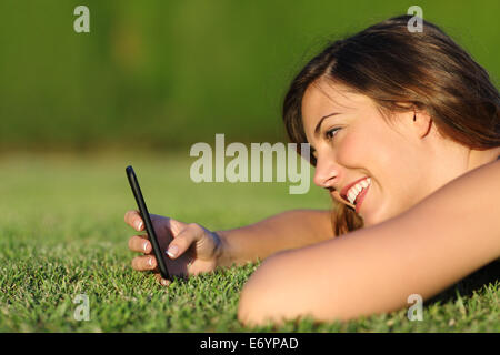 Profil d'une fille drôle en utilisant un téléphone intelligent sur l'herbe d'un parc avec un fond vert Banque D'Images