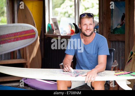 Young male surfer assis dans faire cirer his surfboard Banque D'Images