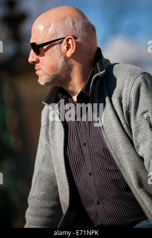 Middle-aged homme chauve en jeans assis dehors au soleil sur un vol de pas lire un journal Banque D'Images