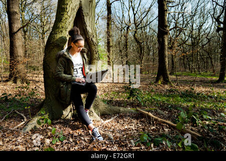 Young mixed race girl s'appuie contre un arbre dans une forêt et se connecte à son ordinateur portable. Banque D'Images