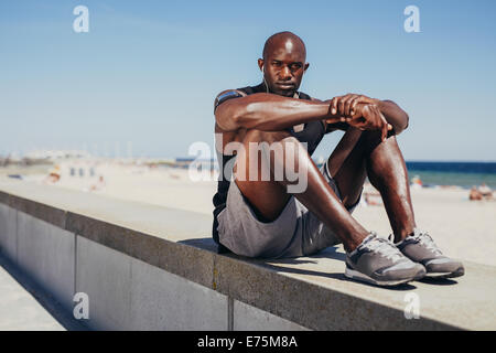 Portrait de monter jeune athlète détente sur sea wall looking at camera. L'Afrique de l'homme musclé se reposant après son entraînement. Banque D'Images