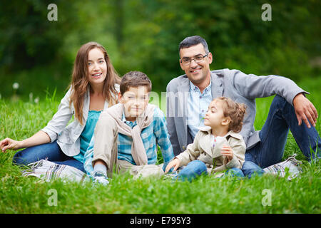 Les membres de la famille heureuse à la caméra à tout en restant assis dans l'herbe verte à l'extérieur Banque D'Images