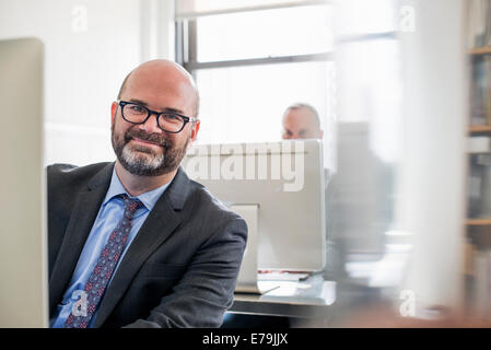 La vie de bureau. Un homme en costume et cravate assis à son bureau à la recherche autour de son ordinateur. Banque D'Images