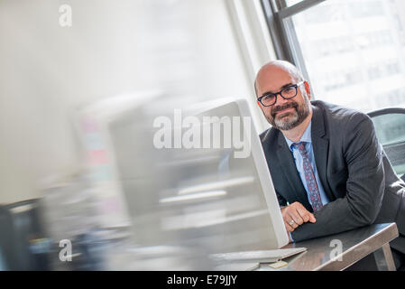 La vie de bureau. Un homme en costume et cravate assis à son bureau à la recherche autour de son ordinateur. Banque D'Images