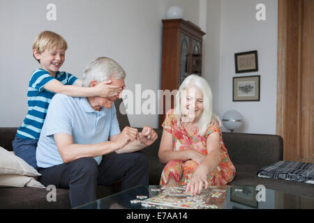 Garçon couvrant les yeux de grand-père alors que la femme l'organisation puzzle à table dans la salle de séjour Banque D'Images