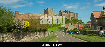 Vue panoramique de l'immense et historique château de Bamburgh sur colline herbeuse avec des chalets de la ville et les gens en premier plan dans le cadre de ciel bleu en Angleterre Banque D'Images