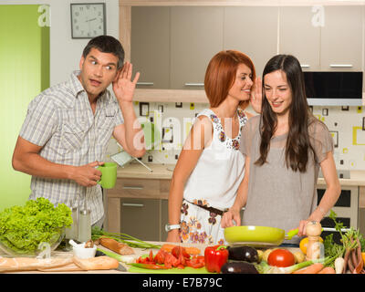 Portrait de deux belles femmes de race blanche debout dans la cuisine cuisine, rire et murmurer quelque chose, avec un homme derrière e Banque D'Images