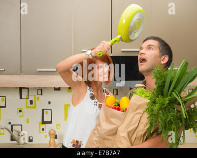 Close-up of young woman frapper surpris choqué l'homme sur la tête avec une poêle à frire, à la fois debout dans la cuisine Banque D'Images