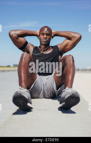 Image du jeune homme fit faire des redressements assis en regardant la caméra. Modèle de remise en forme de l'Afrique de l'exercice en plein air. Banque D'Images