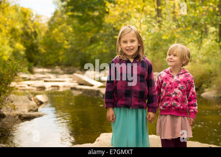 Deux jeunes filles se tenant la main par River dans les rayons du soleil. Fille plus âgée regardant la caméra tandis que les plus jeunes on regarde jusqu'à elle. Banque D'Images