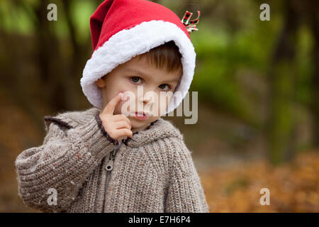 Triste petit garçon dans le parc, avec santa hat, montrant sa joue blessée par la chute, regardant la caméra Banque D'Images