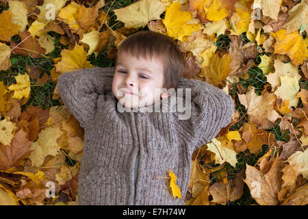 Adorable petit garçon, allongé sur le sol dans le parc, les feuilles autour de lui, souriant à la caméra, en plongée des Banque D'Images