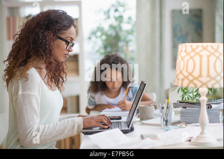 Woman using laptop at home, girl (8-9) à faire leurs devoirs en arrière-plan Banque D'Images