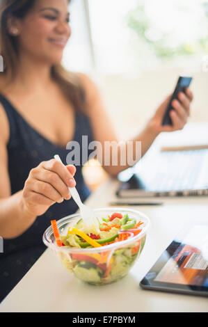 Woman eating salad et à l'aide de mobile phone in office Banque D'Images