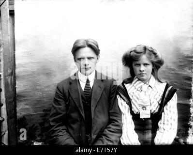 Portrait d'un jeune couple, probablement pris entre 1910 et 1920. L'homme est vêtu d'une cravate, tandis que la femme porte une robe rayée. La photo capture un moment solennel du couple assis ensemble, dégageant un charme vintage. Banque D'Images