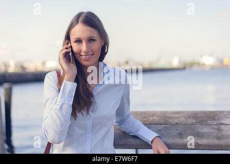Jolie femme de prendre un appel sur son téléphone portable à l'extérieur debout contre un arrière-plan de la rivière smiling at the camera Banque D'Images