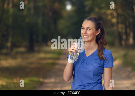 Jeune femme élancée fit boire de l'eau embouteillée dans les vêtements de sport alors qu'elle s'arrête sur une piste forestière à réhydrater pendant un run de formation Banque D'Images