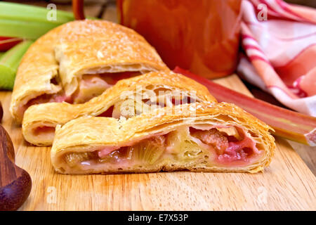 Strudel aux pommes avec de la rhubarbe, couteau, tasse, serviette, tiges de rhubarbe sur le fond de panneaux de bois Banque D'Images