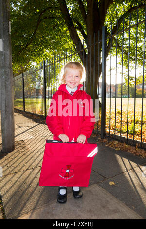 Un enfant de 4 ans / fille dans son nouvel uniforme rouge aller à / sur le chemin de sa classe d'accueil à l'enfant de l'État l'école primaire. UK. Banque D'Images