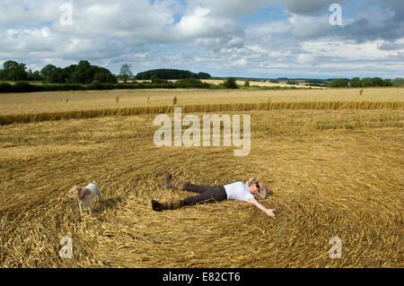 Un agriculteur en étant couché sur le dos dans les chaumes d'un champ de récolte fraîchement coupé de la création d'un motif dans la paille. Banque D'Images