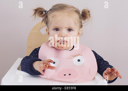 Baby Girl sitting in chaise haute avec la nourriture enduite sur son visage et ses mains Banque D'Images