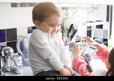 Tout-petit garçon assis sur le comptoir de la cuisine, souriant comme sœur atteint pour candy jar Banque D'Images