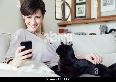 Femme assise sur un canapé avec chat sur ses genoux, using smartphone Banque D'Images