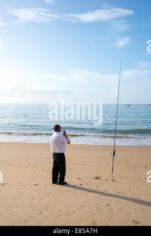 Homme debout sur la plage de cascais en pantalon noir et chemise blanche avec la canne à pêche, à quelques mètres de la fish restaurant il travaille Banque D'Images