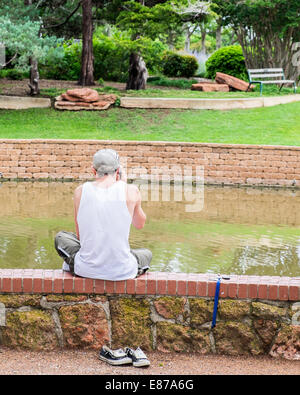 Un jeune homme de race blanche parlant sur un téléphone mobile se trouve sur un mur dans le parc Will Rogers, Oklahoma City, Oklahoma, USA. Banque D'Images