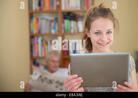 Portrait de grand-père lisant le journal, petite-fille à l'aide de digital tablet in living room, smiling Banque D'Images