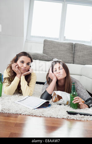 Ennuyer deux amies situées côte à côte sur un tapis à la maison à regarder la télévision Banque D'Images