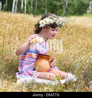 Petite fille est assise sur l'herbe jaune avec verseuse et le pain dans la main outdoor Banque D'Images