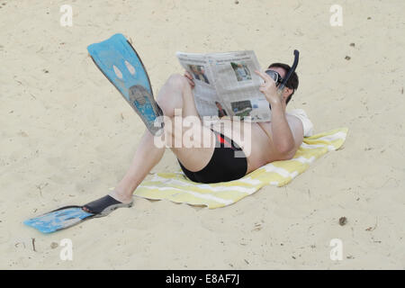 L'homme à palmes et masque de plongée avec tuba jette sur le sable et lit journal Banque D'Images