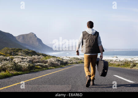 Vue arrière du jeune homme avec l'étui à guitare marche sur route côtière, Cape Town, Western Cape, Afrique du Sud Banque D'Images