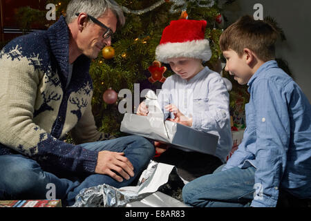 Père sur l'ouverture du plancher salon des cadeaux de noël avec deux fils Banque D'Images