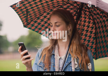 Jeune femme avec parapluie texting on smartphone in park Banque D'Images