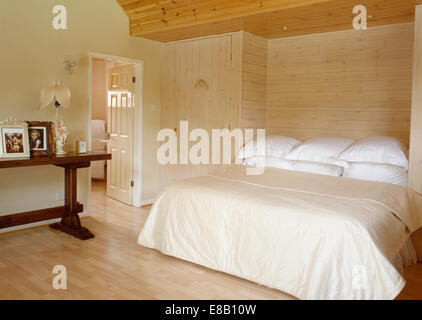 Derrière un mur lambrissé de bois clair lit avec oreillers et couette blanc crème en cottage moderne chambre avec parquet au sol Banque D'Images