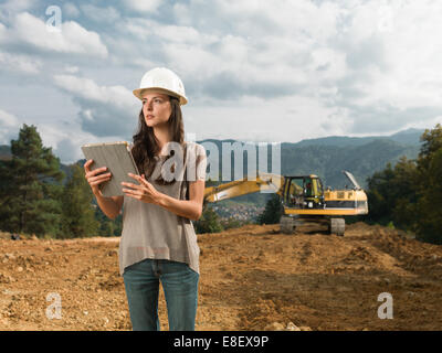 Portrait of young caucasian female engineer debout sur site de construction, holding digital tablet, looking away Banque D'Images