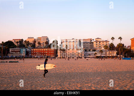 Un surfeur à marcher le long de la plage de Santa Monica, en Californie, au coucher du soleil Banque D'Images