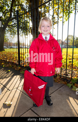 Un enfant de 4 ans / fille dans son nouvel uniforme rouge aller à / sur le chemin de sa classe d'accueil à l'enfant de l'État l'école primaire. UK. Banque D'Images