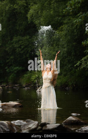 Corbeille la robe : un 'bride' de porter sa robe de mariage debout dans une rivière jetant de l'eau dans l'air Banque D'Images