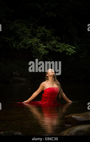 Une femme aux cheveux blonds portant une fille robe de bal robe rouge profond jusqu'à sa taille dans l'eau dans une rivière avec ses yeux fermés Banque D'Images