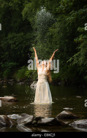 Corbeille la robe : un 'bride' de porter sa robe de mariage debout dans une rivière jetant de l'eau dans l'air Banque D'Images