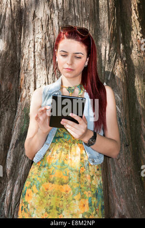 Belle jeune fille leaning ona arborescence à l'aide d'ipad Banque D'Images