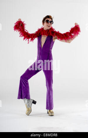 Dans l'homme fancy dress costume de comédie en 1970 camp flamboyant costume avec disco purple catsuit, plate-forme chaussures & feather bower Banque D'Images