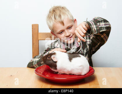 Enfant garçon blond assis à la table à manger avec des couverts cochon (viande crue) assis sur une plaque rouge Banque D'Images