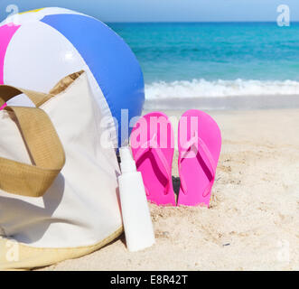 Sac de plage et boule colorée avec la crème solaire et des tongs sur le sable près de la ligne d'eau à la plage Banque D'Images