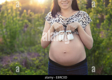 Close up of méconnaissable femme enceinte avec petites chaussures bébé sur son ventre au lever de l'arrière-plan Banque D'Images