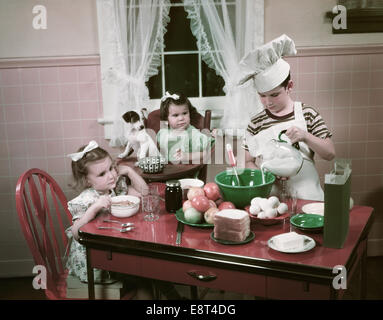 Années 1940 Années 1950 ENFANTS DEUX FILLES À TABLE DE CUISINE AVEC CHIOT ET BOY WEARING CUISINE CHEFS TOQUE Banque D'Images